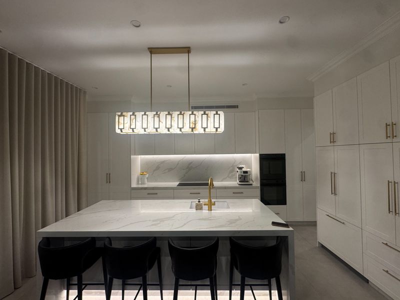 A long marble island with black barstools beneath a pendant light, gold tap and full-height cabinetry