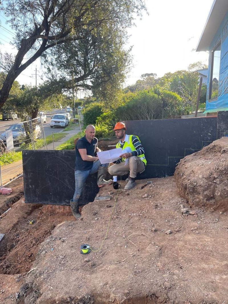 Two tradespeople going through plans on site beside a tanked retaining wall, before excavation is backfilled