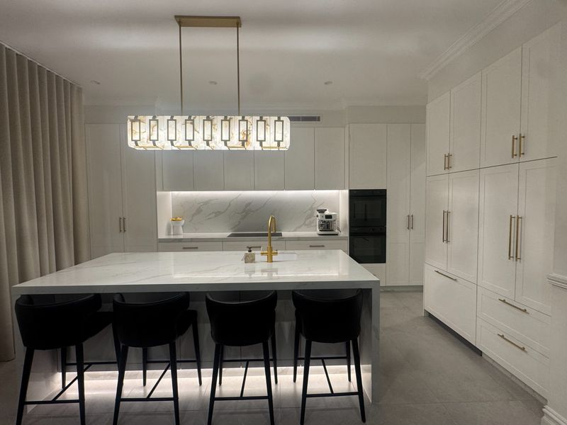 A long marble island with black barstools sits beneath a modern pendant light, with an oven and cabinetry along the back wall.